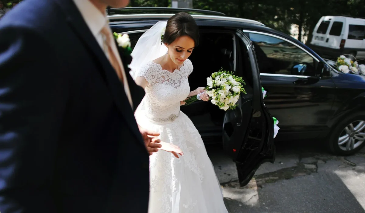 Elegant bride in a white wedding gown and veil steps gracefully out of a luxury black chauffeur-driven car, holding a bouquet of flowers. A groom in a suit stands nearby to assist her.