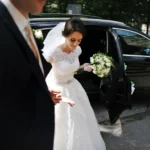 Elegant bride in a white wedding gown and veil steps gracefully out of a luxury black chauffeur-driven car, holding a bouquet of flowers. A groom in a suit stands nearby to assist her.