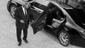Professional chauffeur in a suit opens the rear door of a luxury black Mercedes saloon for a passenger seated inside. Represents MCR EXEC’s executive and corporate chauffeur services.