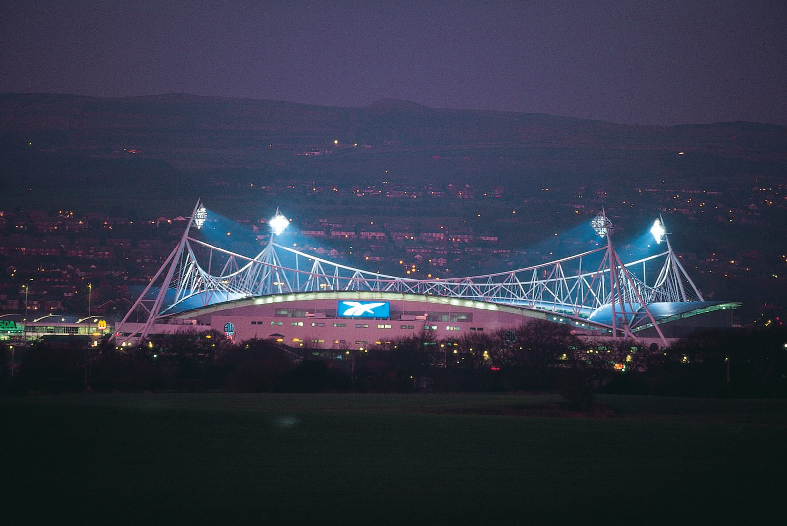 Night view of the illuminated Bolton Arena stadium with its distinctive white structural roof supports lit up against the evening sky. Represents MCR EXEC’s Bolton Arena Chauffeur Services for events and matches.