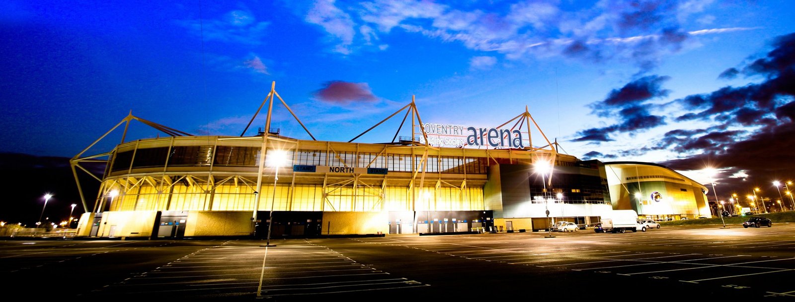 Evening view of the illuminated Coventry Building Society Arena with striking yellow and gold lighting against a twilight sky. The venue’s exterior signage is clearly visible, representing MCR EXEC’s arena chauffeur services.