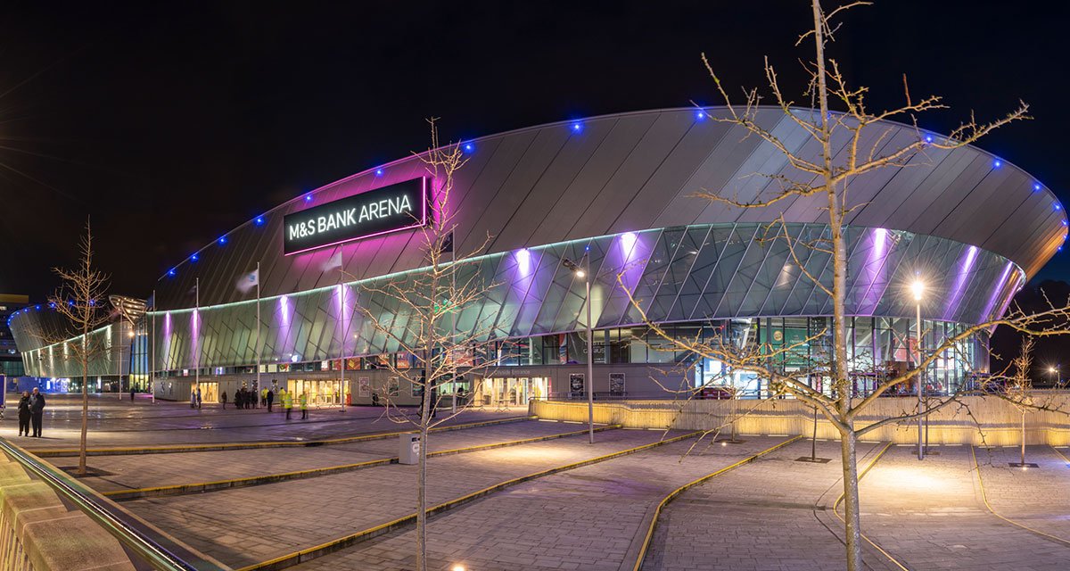 Night view of Liverpool’s M&S Bank Arena with purple and blue exterior lighting, modern architecture, and people entering the venue. Represents MCR EXEC’s chauffeur services for arena events.