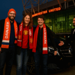 Three Danish Manchester United fans in red scarves and jackets pose outside Old Trafford after arriving in a black Mercedes MPV. A suited chauffeur greets them.