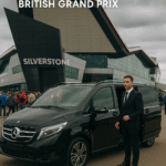 Professional chauffeur stands beside a black Mercedes V-Class outside Silverstone Circuit with a crowd in the background and bold “RESERVE NOW” CTA for MCR EXEC service from Liverpool.