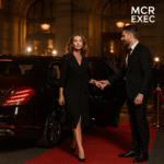 A woman in an elegant black dress steps out of a Mercedes S-Class onto the red carpet outside a Manchester venue, assisted by a professional chauffeur.