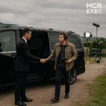 A film director wearing headphones shakes hands with a chauffeur beside a black Mercedes V-Class on a rural film set outside Manchester.