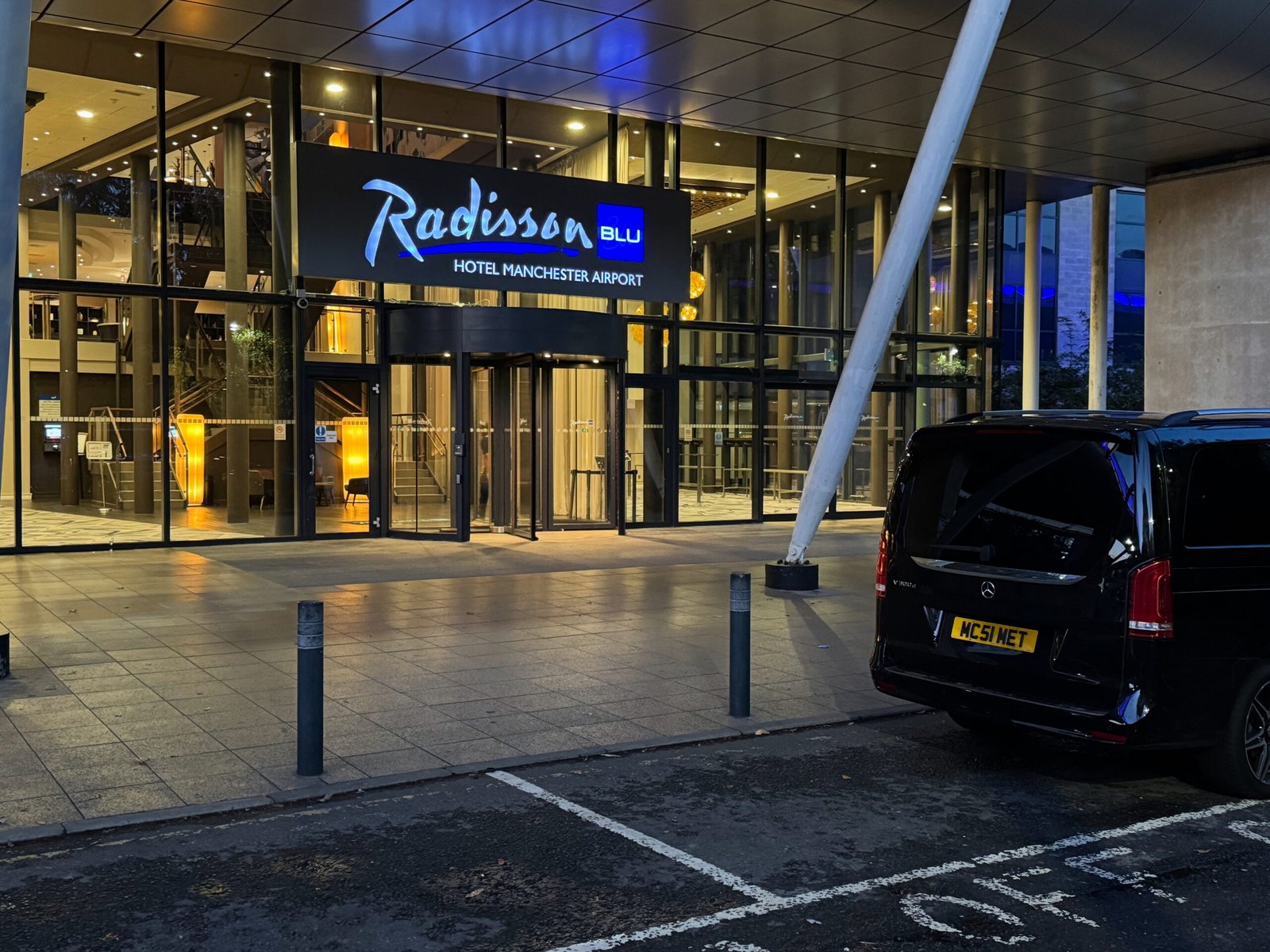 Black Mercedes V-Class chauffeur vehicle parked outside the Radisson Blu Hotel Manchester Airport at dusk, providing executive transport services for hotel guests.