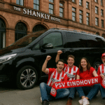 PSV Eindhoven Dutch football fans outside The Shankly Hotel in Liverpool, celebrating with a red-and-white scarf beside a black Mercedes V-Class chauffeur car, MCR EXEC branding included.