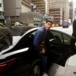 Professional chauffeur services in Manchester with a black Mercedes E-Class by MCR EXEC, chauffeur adjusting tie before client pickup outside a city hotel.