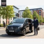 Black Mercedes V-Class with MCR-EXEC plate in Hulme, Manchester, chauffeur assisting a passenger beside modern buildings near Birley Fields Campus.
