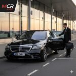 Irlam Airport Chauffeur Transfers by MCR-EXEC — a black 2025 Mercedes S-Class with MCR-EXEC number plates positioned at Manchester Airport Terminal 2 during early sunrise. A professional chauffeur in a tailored suit (no hat or gloves) assists a business traveller with rolling luggage at the curb. The glass façade of the terminal reflects the dawn light. The MCR EXEC logo is displayed in the top-left corner. This image represents premium airport chauffeur service from Irlam, connecting executives and families to Manchester Airport with professionalism, comfort, and reliability