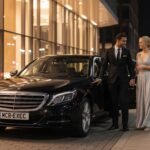 Overnight chauffeur hire for events using a black Mercedes S-Class with MCR-EXEC number plate, professionally dressed chauffeur escorting an evening event couple outside a modern city venue