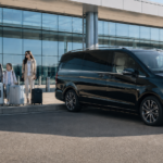 Family with luggage meeting a chauffeur beside a black Mercedes V-Class at an airport terminal.